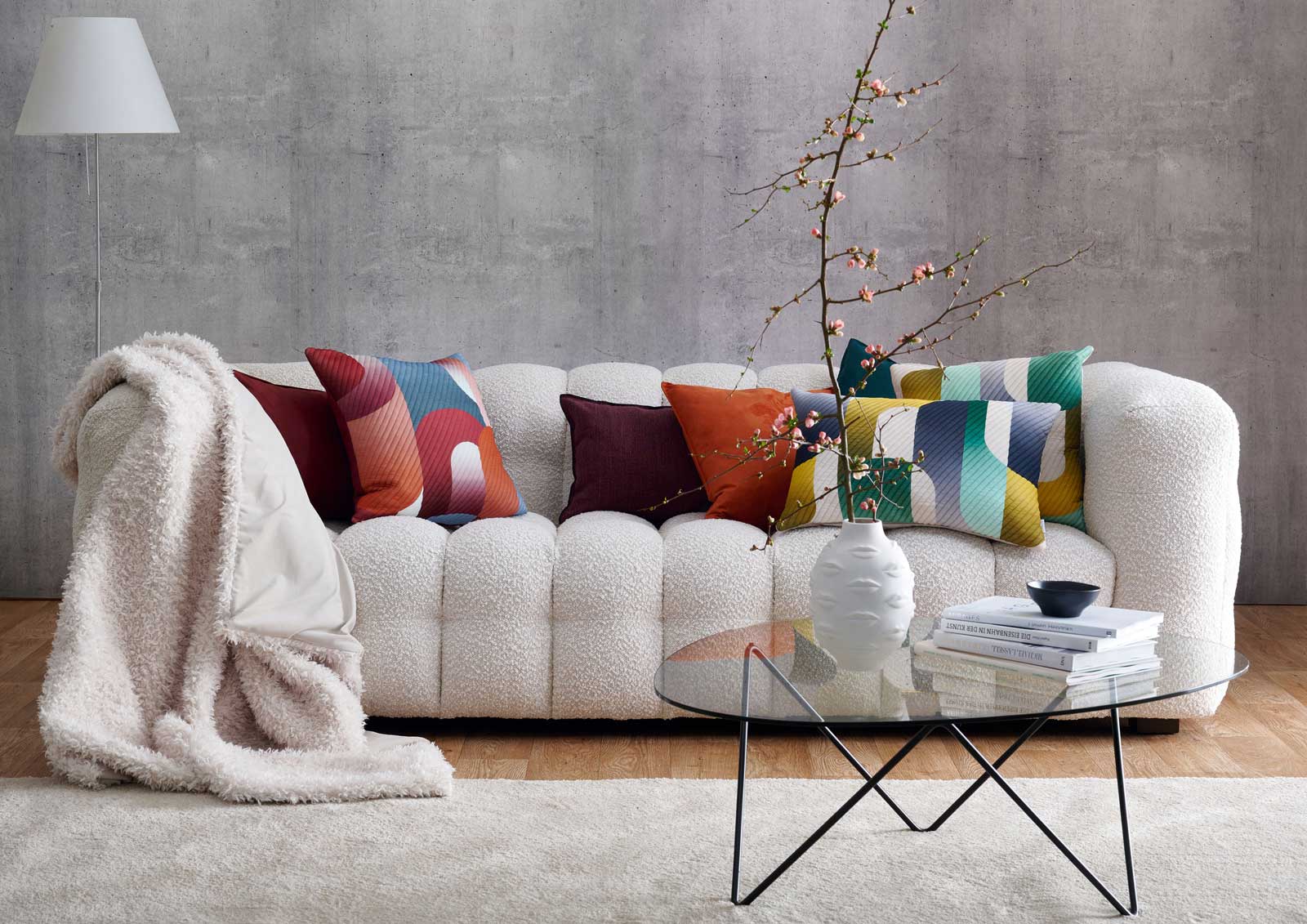 wollweisses Sofa mit Felldecke und bunten Kissen - davor ein kleiner runder Tisch mit Vase und B&uuml;chern