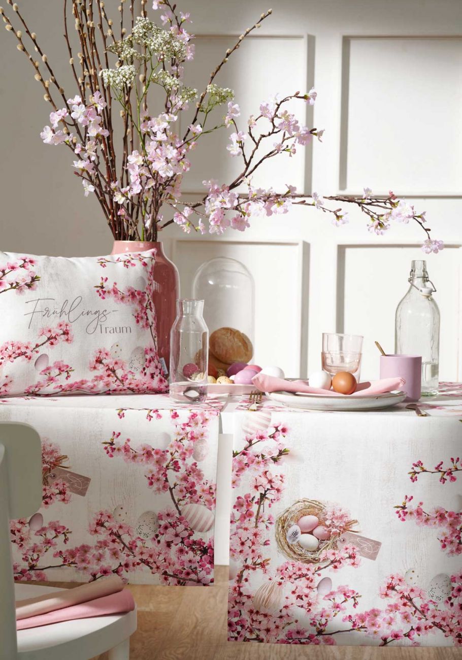 Tischläufer und Kissen mit Ostermotiven (Blumen und Ostereier) weiß mit rosa Dekor darauf eine Vase mit Blüten und Geschirr
